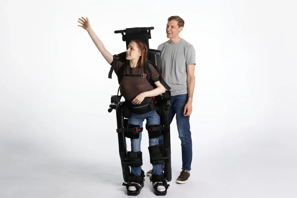 Une utilisatrice debout dans l'exosquelette Wandercraft Eve, la main levée, son accompagnant derrière elle